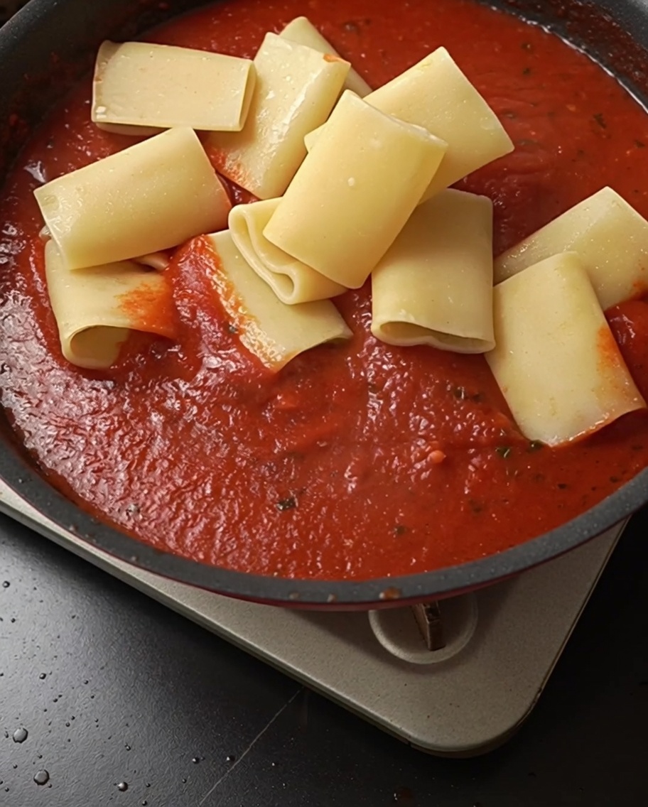 Cooked pasta tubes being added to a pan filled with vibrant red tomato sauce.