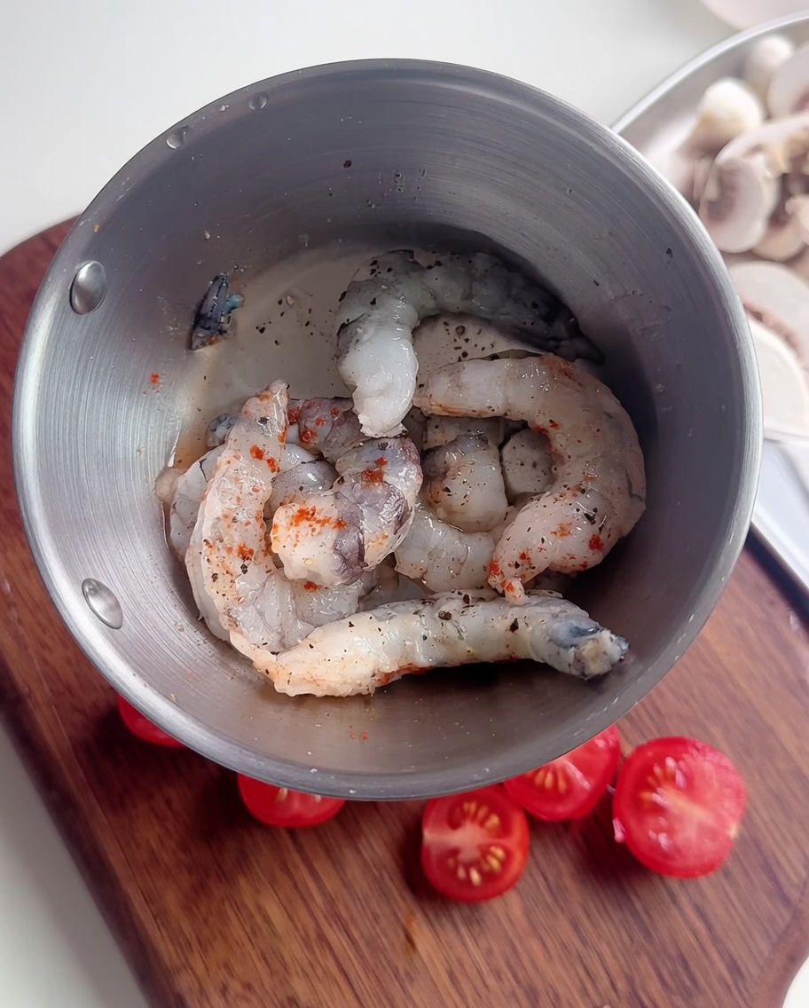 A close-up of raw shrimp in a metal bowl seasoned with black pepper, red chili powder, and a thin starch slurry.