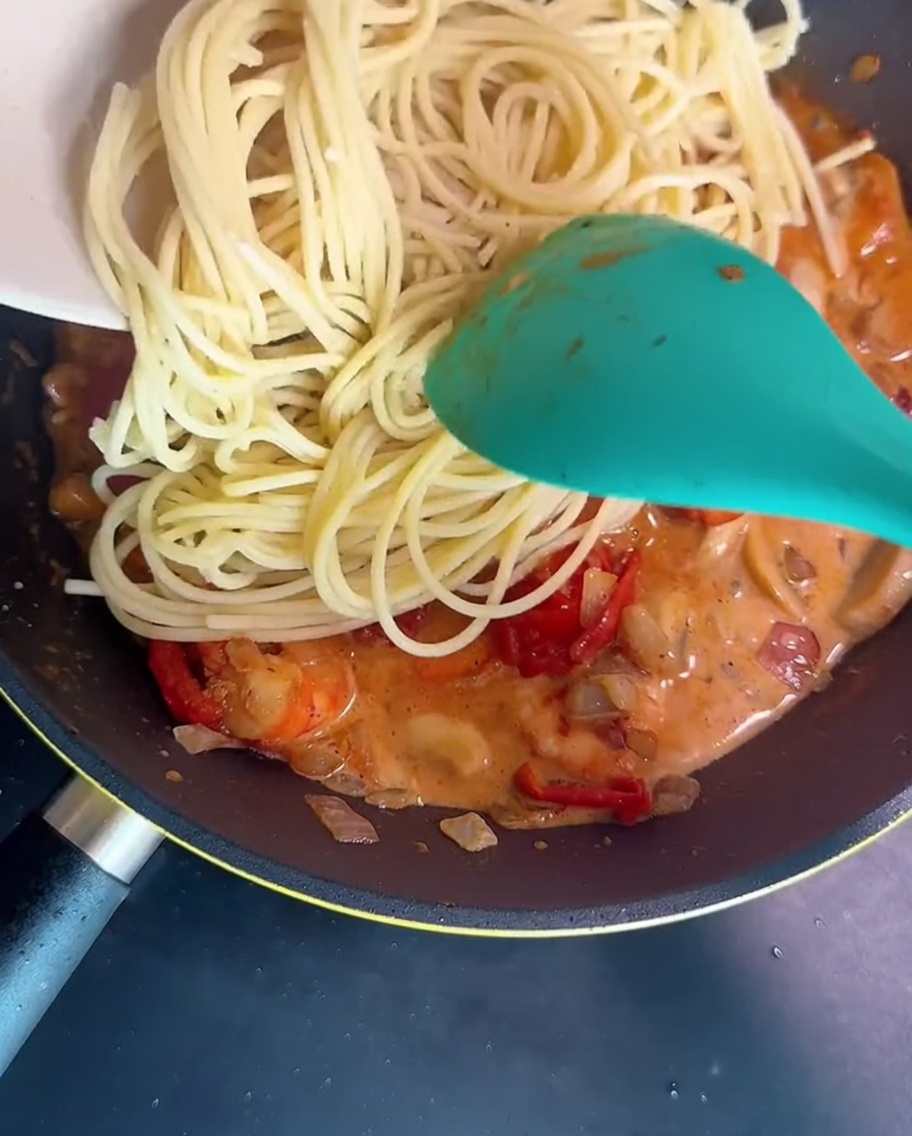 Spaghetti being tossed in a frying pan with a creamy shrimp and mushroom sauce using a green spatula.