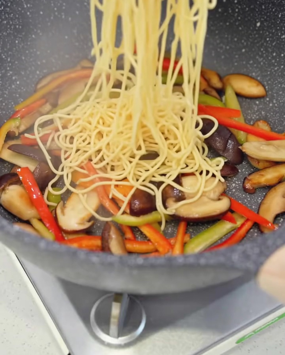 Cooked spaghetti strands being dropped into a pan filled with sautéed mushrooms and colorful bell peppers.