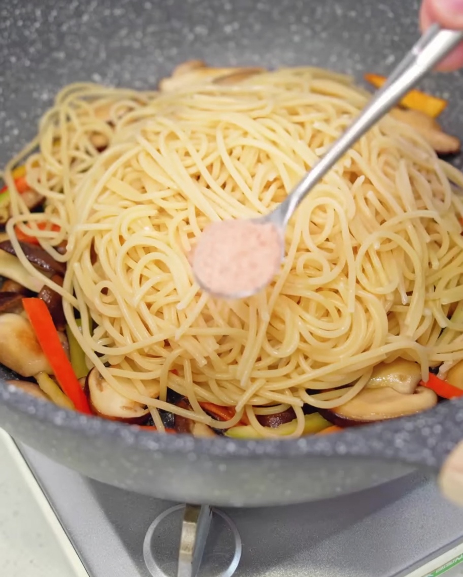 A small spoonful of pink salt being sprinkled over the spaghetti in the cooking pan.