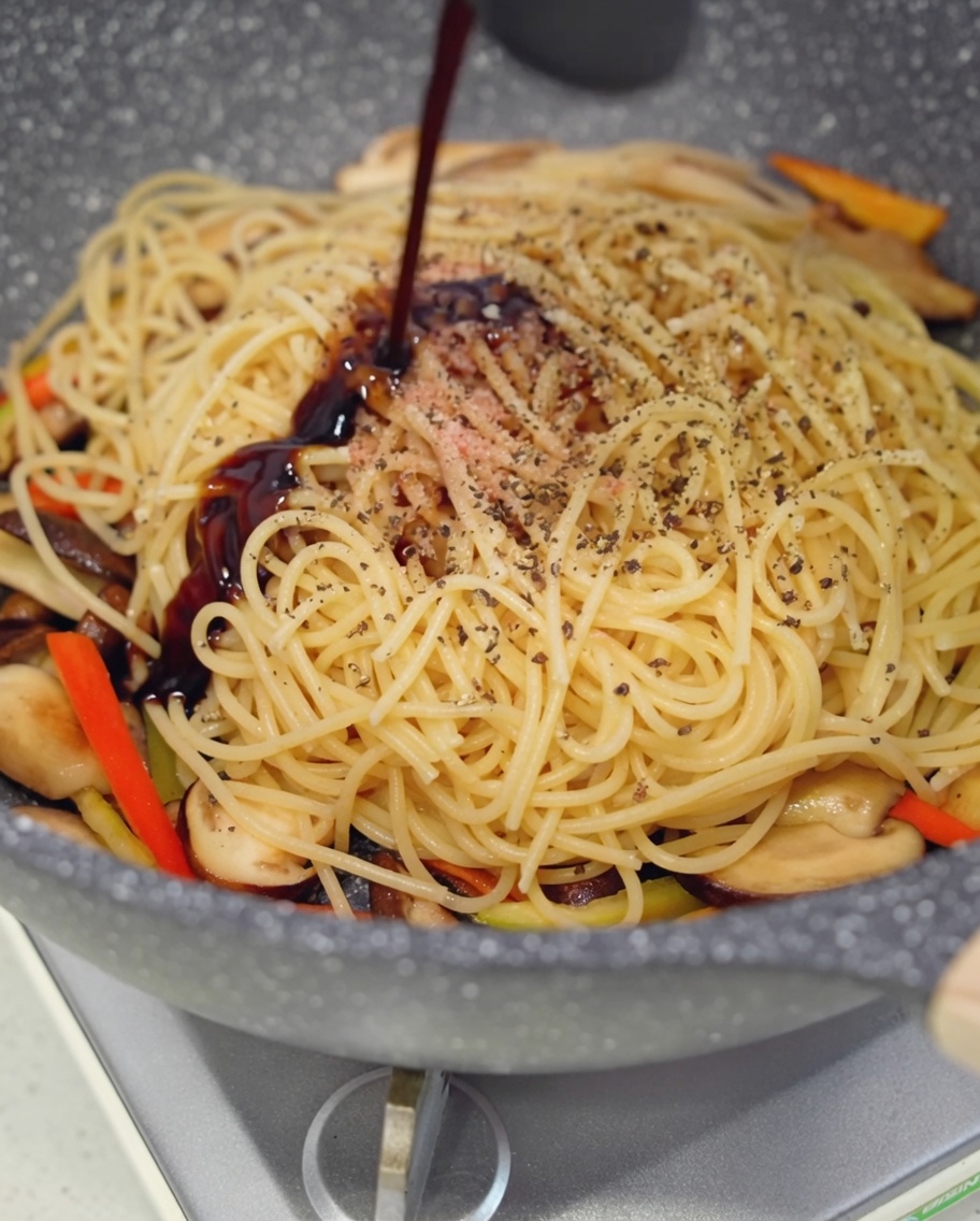 Dark vegan oyster sauce being drizzled onto the spaghetti and mushroom mixture in the pan.