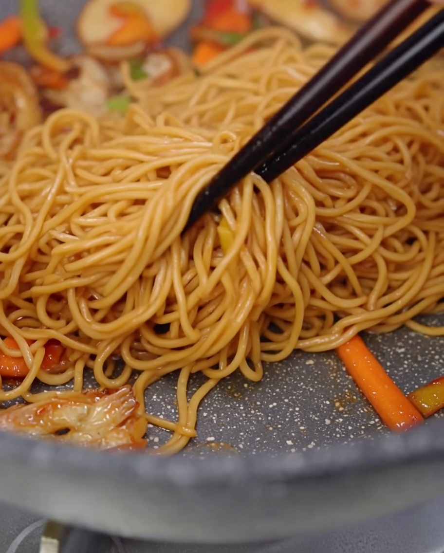 A close-up of spaghetti, sliced mushrooms, and colorful bell pepper strips being tossed together in a pan using chopsticks.