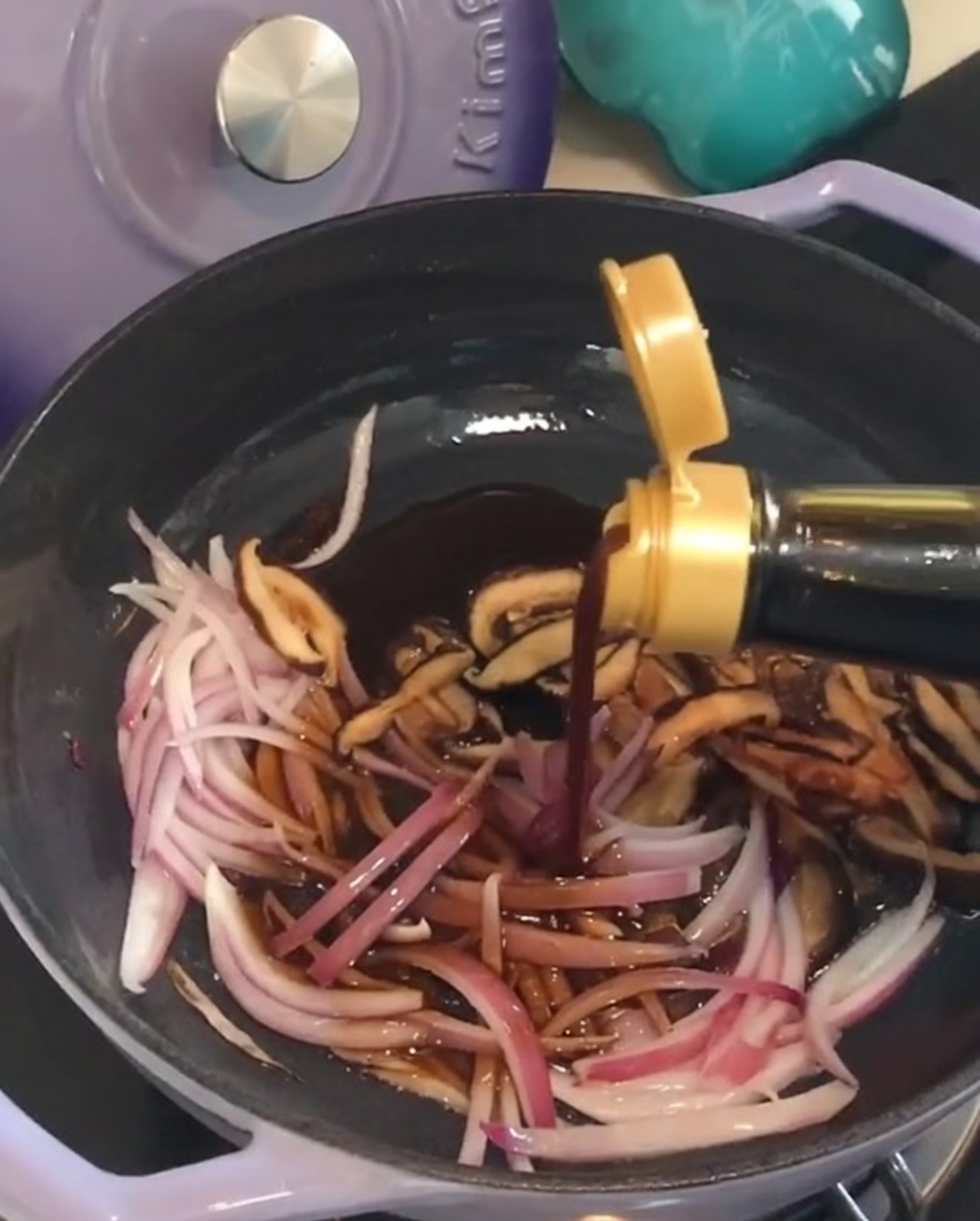 Pouring dark sauce from a bottle into a pan with sautéed red onions and shiitake mushrooms.