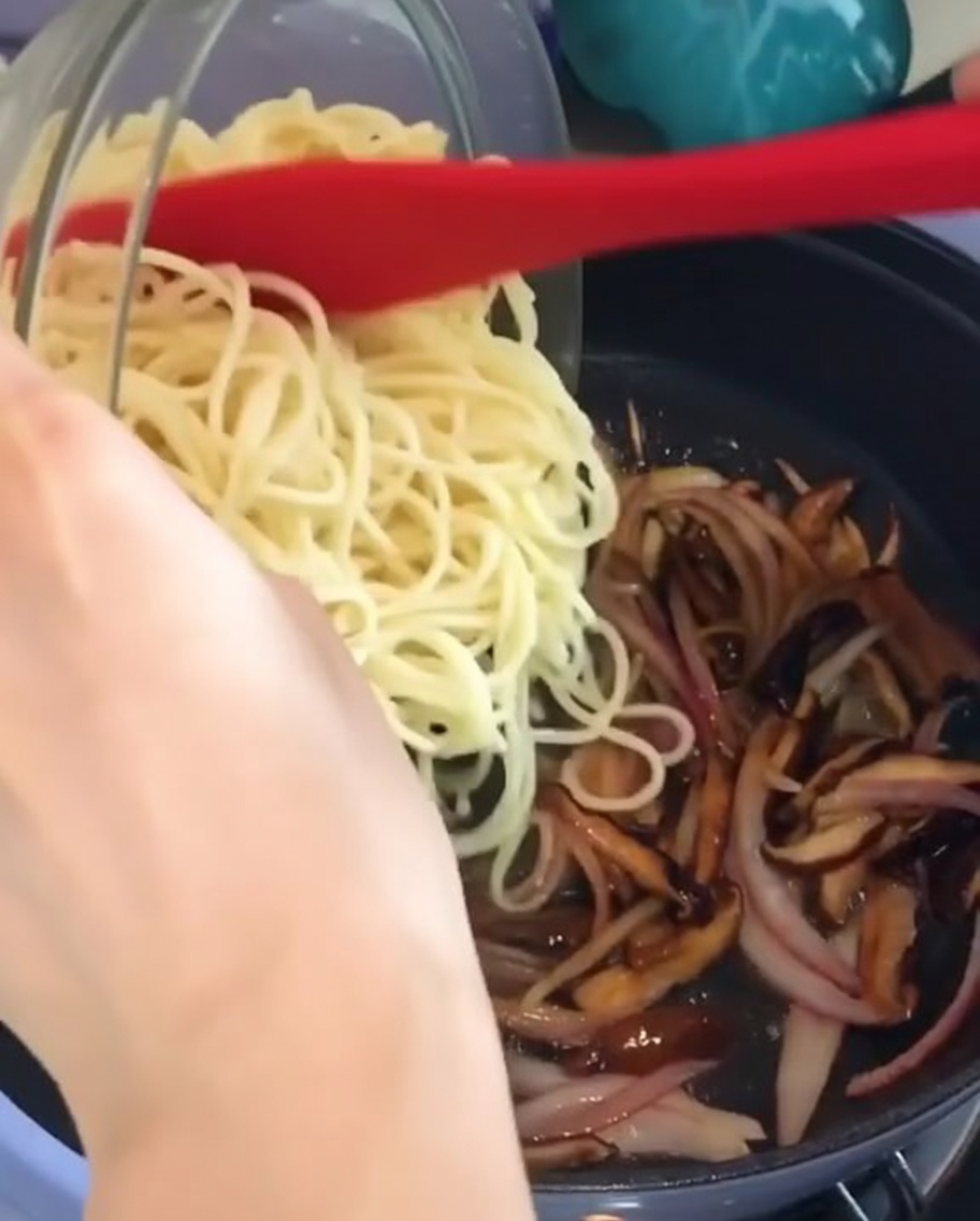 Cooked spaghetti being added from a glass bowl into a pan of cooked onions and mushrooms.