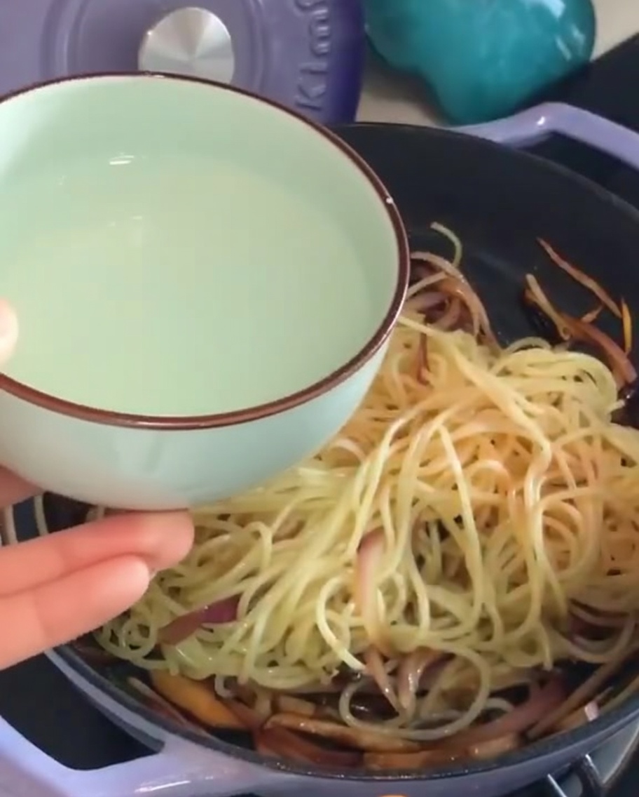 Pouring a small bowl of starchy pasta water into a pan of spaghetti and vegetables.
