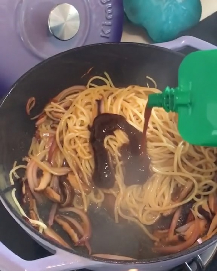 Squeezing black pepper sauce from a green packet into a pan of stir-fried spaghetti.