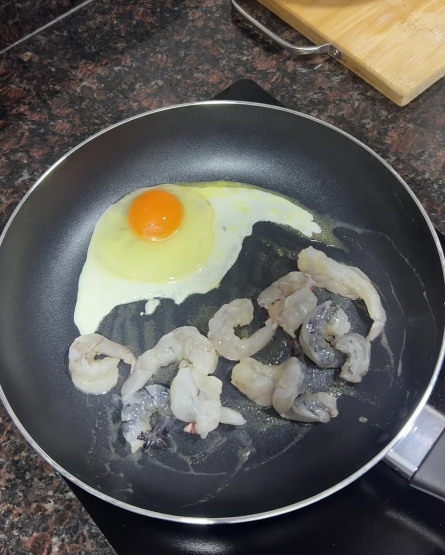 Raw shrimp browning in a pan next to a frying egg.