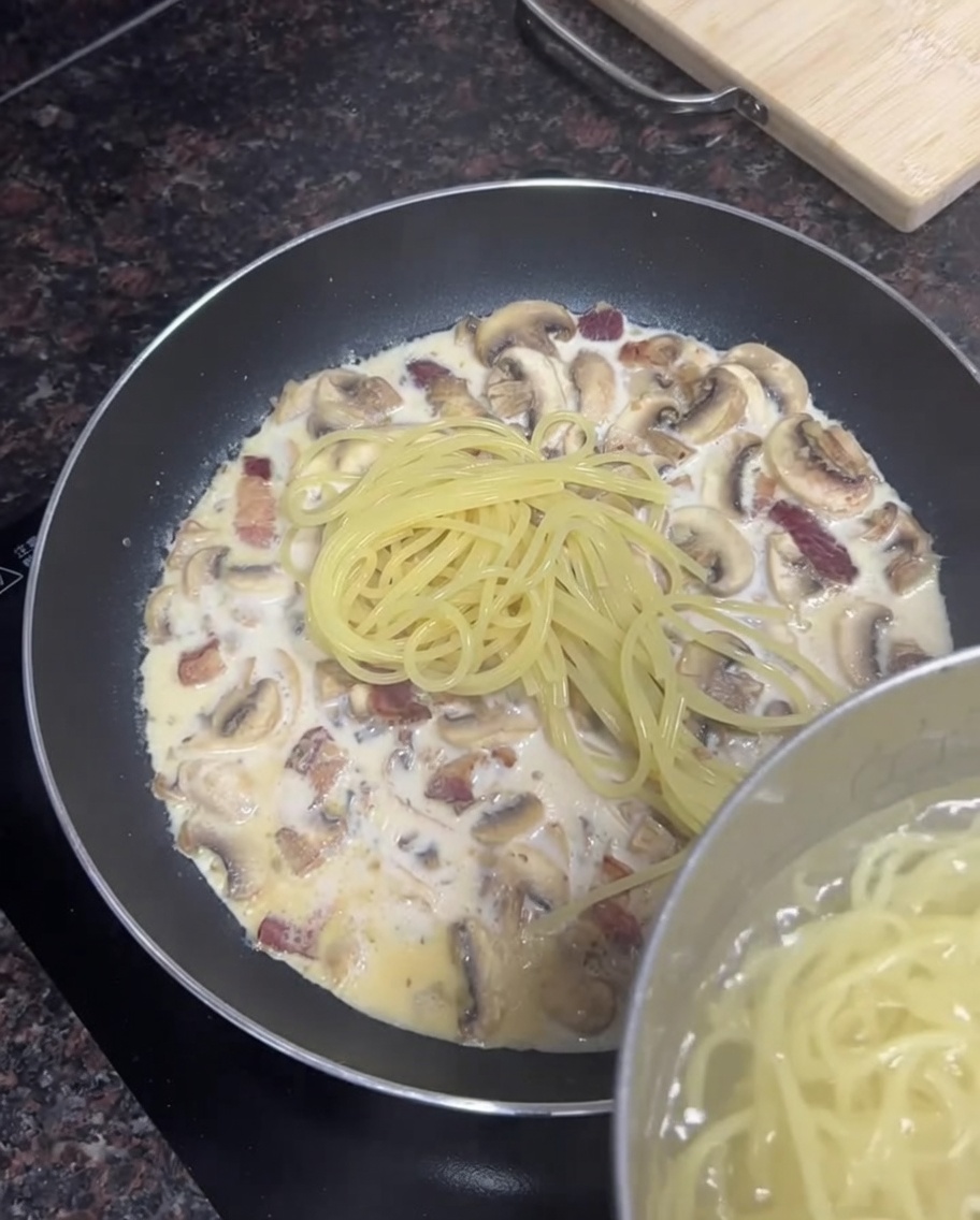 Cooked spaghetti noodles being added from a pot into a frying pan filled with creamy bacon and mushroom sauce.