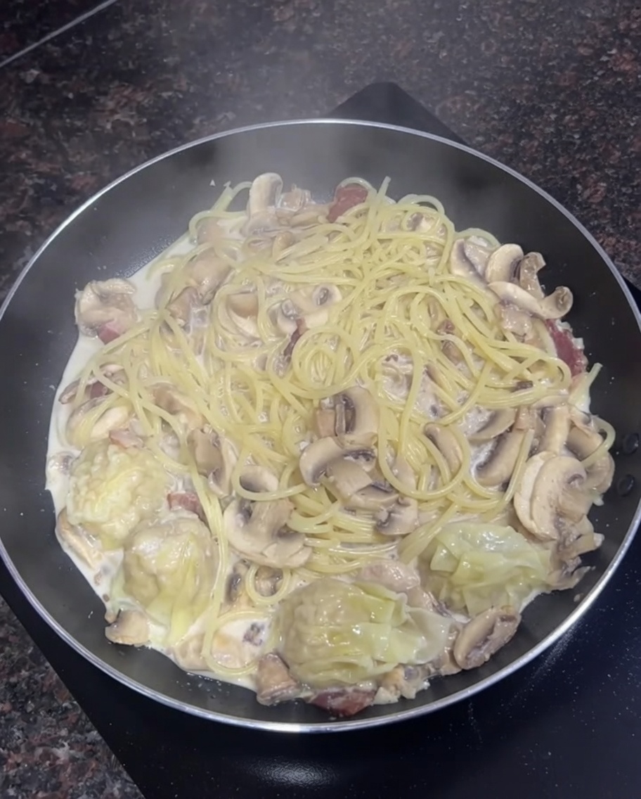 Frying pan filled with creamy spaghetti, sliced mushrooms, and a few cooked wontons simmering together.