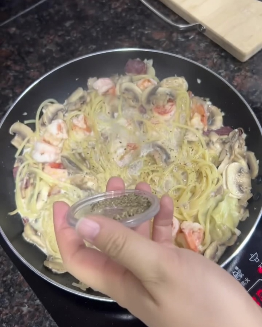 A hand sprinkling fresh black pepper from a small container over a pan of creamy pasta, shrimp, and mushrooms.