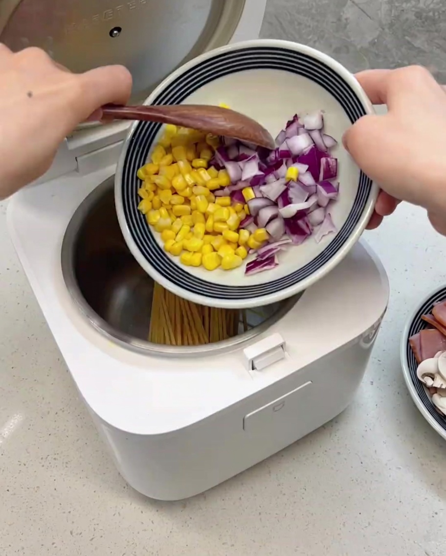 A variety of ingredients including corn, red onion, ham, and mushrooms resting on top of the raw spaghetti in the rice cooker pot.