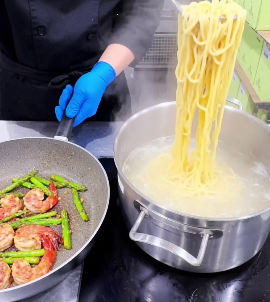 Pasta being added into a skillet containing cooked shrimp, asparagus, and garlic.