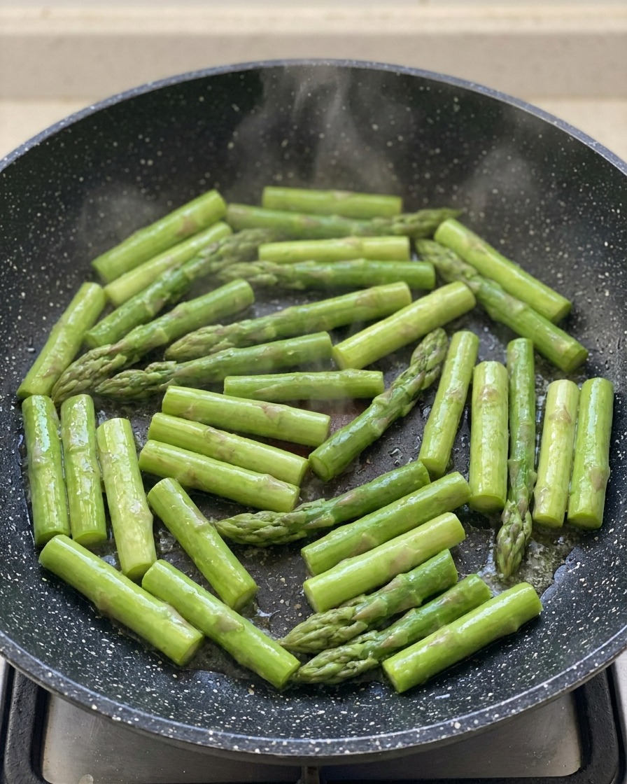 Bright green sliced asparagus spears sizzling in a dark speckled frying pan.