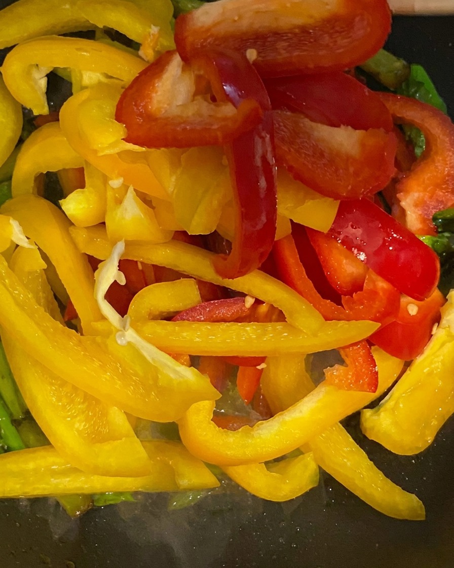 Vibrant yellow and red bell pepper strips being added to a pan with green asparagus.
