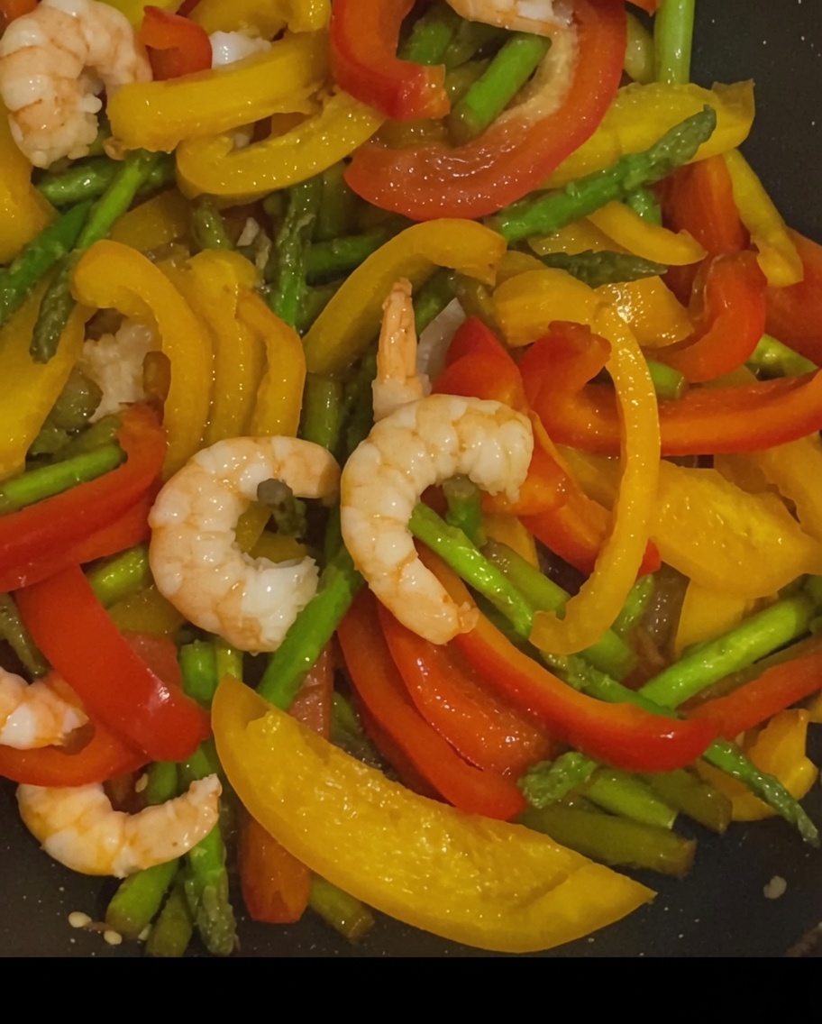 Stir-fried shrimp, yellow and red bell peppers, and asparagus seasoned with specks of black pepper in a hot pan.