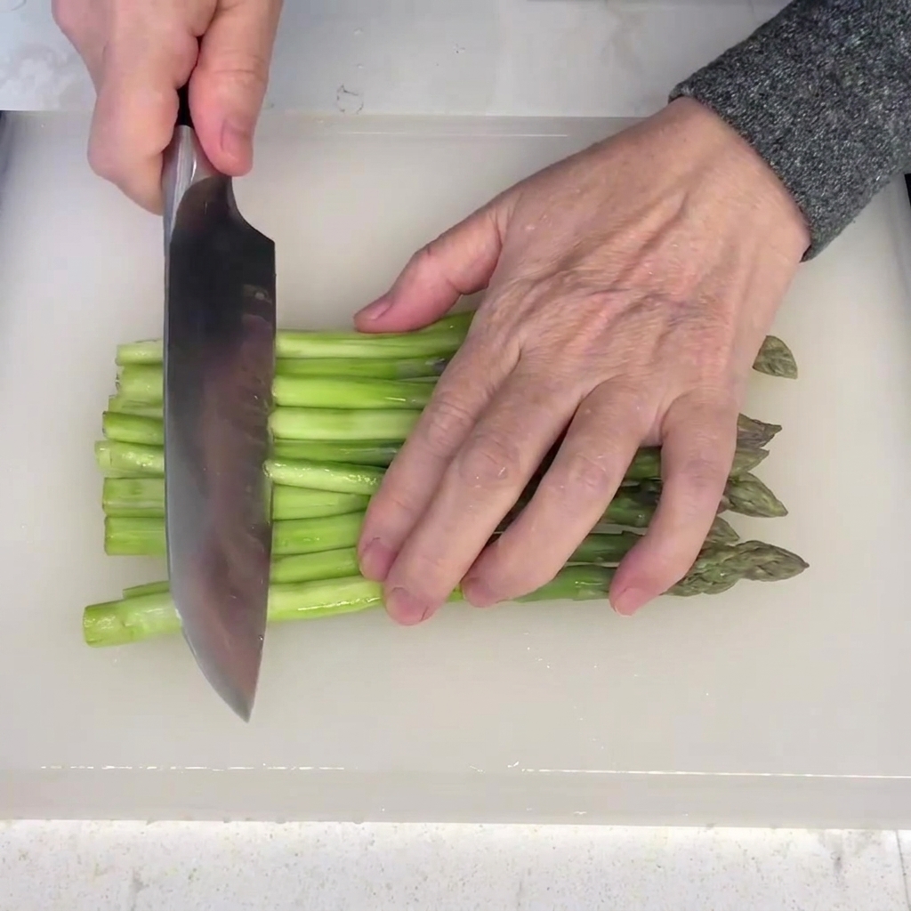 Peeled asparagus stalks being cut into uniform segments on a white cutting board.