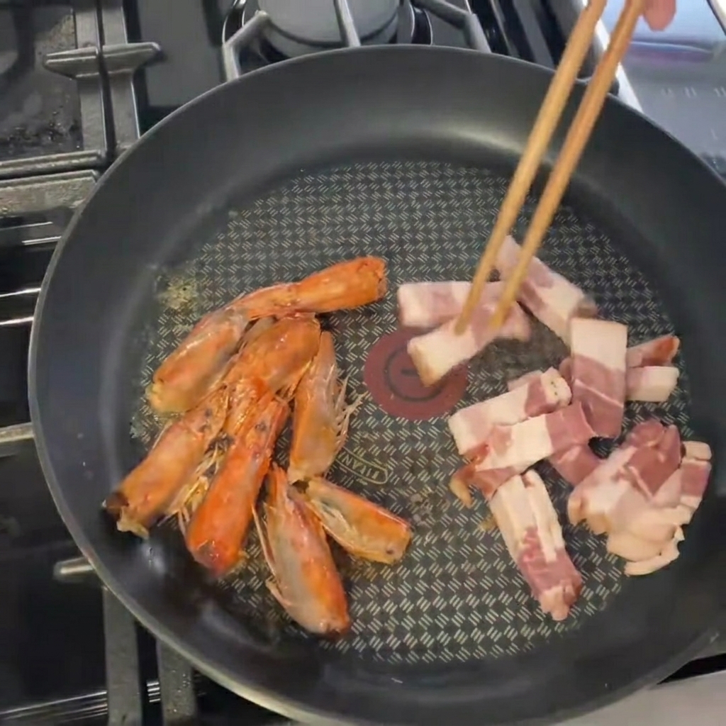 Pieces of sliced bacon being dropped into a pan already containing sautéed shrimp heads.