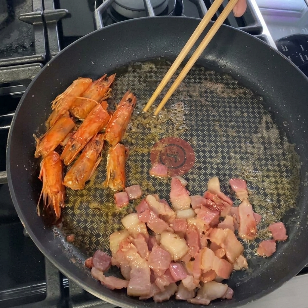 Large, peeled shrimp being placed into the pan alongside bacon and shrimp heads.