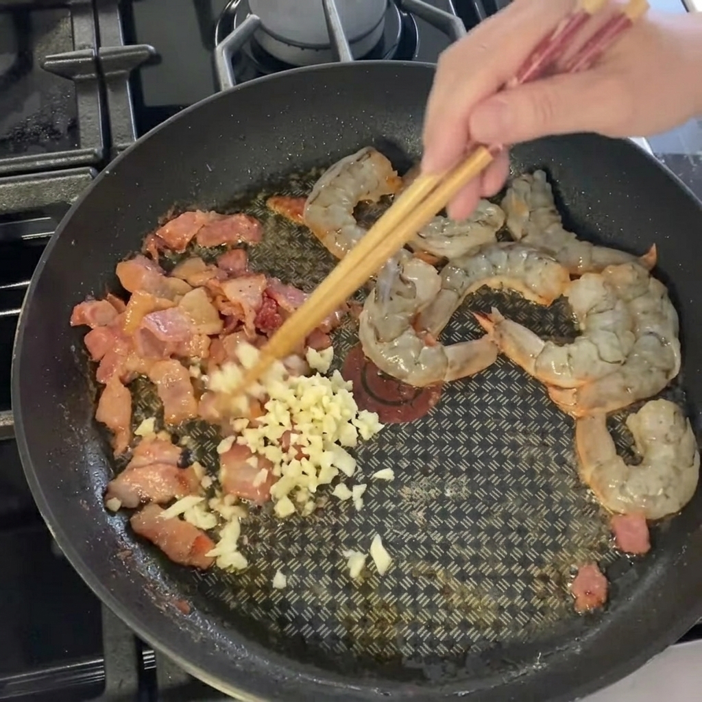 Finely minced garlic being scattered over the shrimp and bacon mixture in the pan.