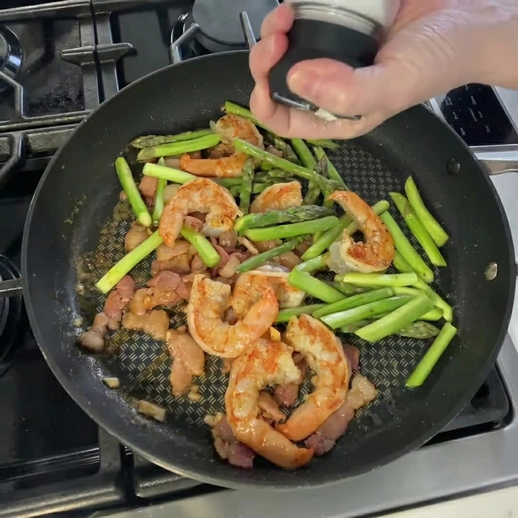Hand seasoning the shrimp and asparagus mixture in the pan with salt and pepper.