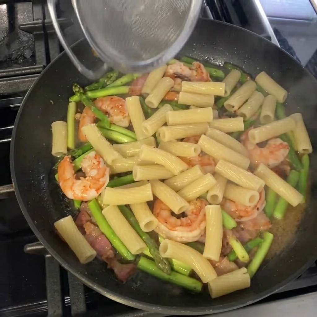 Cooked rigatoni being tossed into the pan with the asparagus and shrimp, ready for final seasoning and garnishing.
