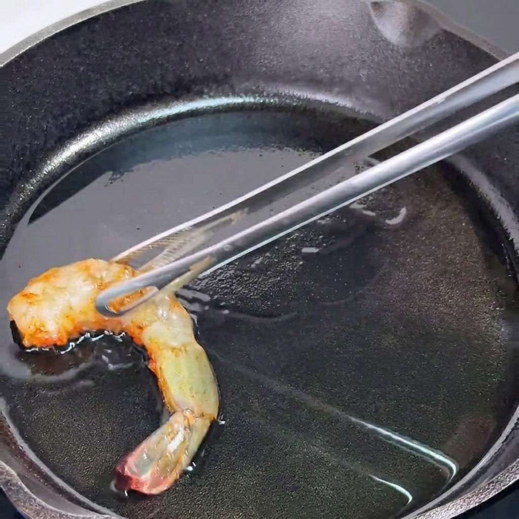 Metal tongs holding a partially cooked, orange-hued shrimp over a hot, oiled cast iron skillet.