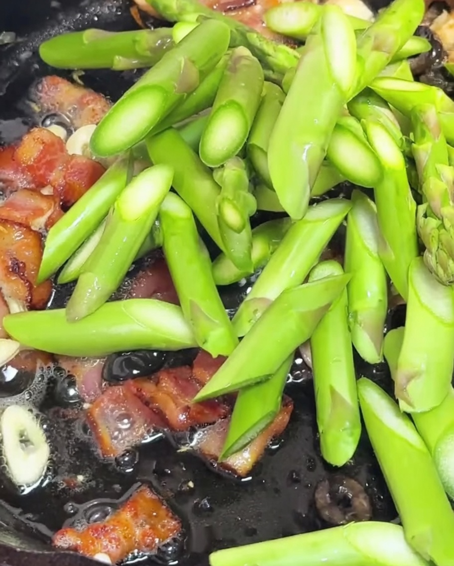 Asparagus pieces and halved cherry tomatoes being sautéed in a pan with bacon and garlic.