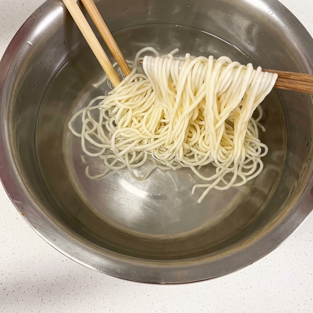 Cooked noodles being held up with chopsticks over a bowl of cold water.