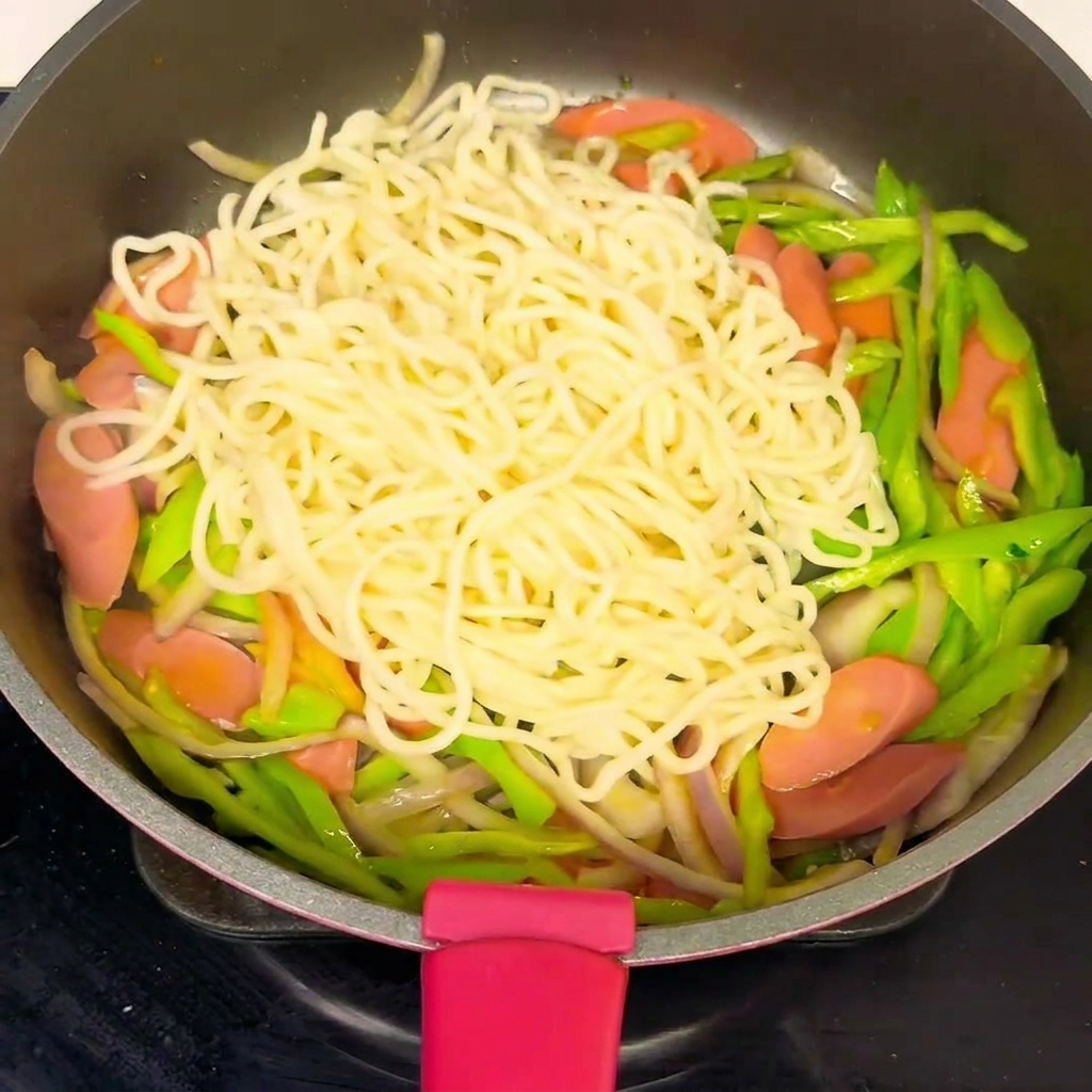 A pan filled with noodles and vegetables, with a sauce being poured over the mixture.