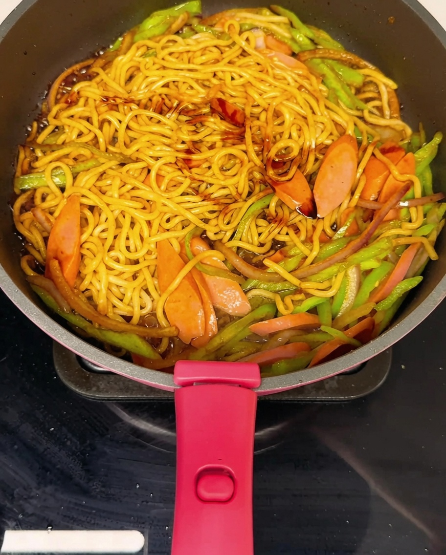 Stir-fried noodles in a pan being seasoned with freshly ground black pepper.