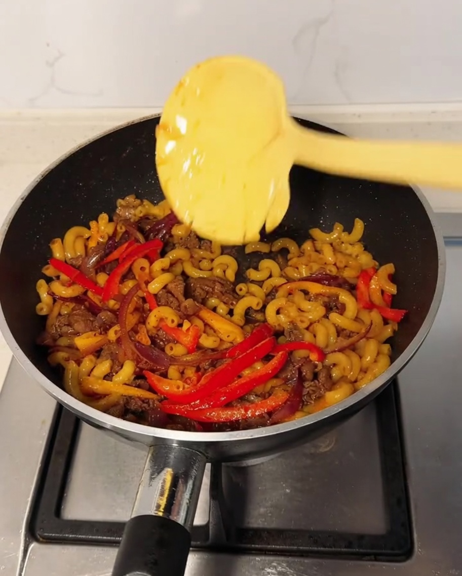 A slice of cheese sits on top of the macaroni mixture in a wok with black pepper powder being sprinkled over it.