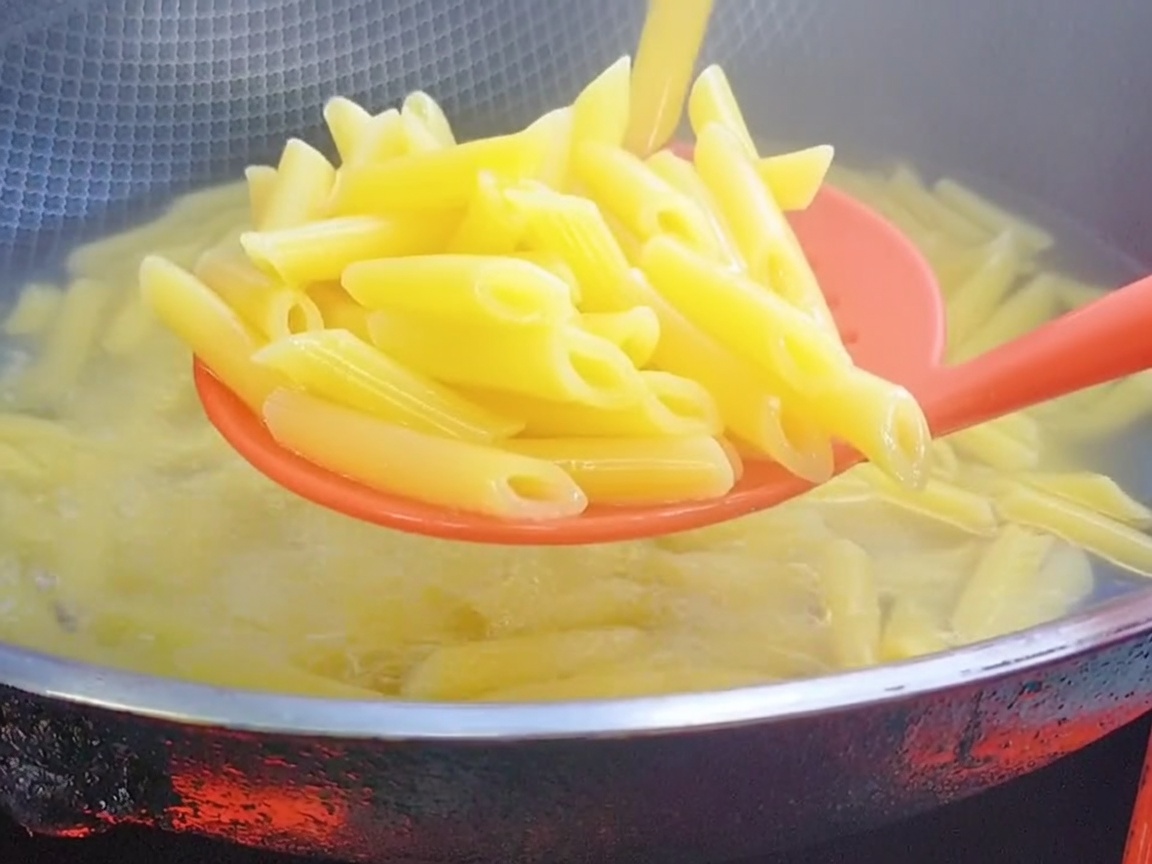Red silicone slotted spoon lifting cooked penne pasta from the boiling water.