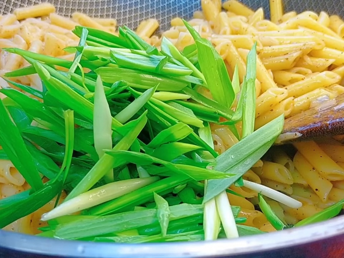 Fresh green garlic sprouts being tossed into a wok filled with penne pasta and black pepper sauce.