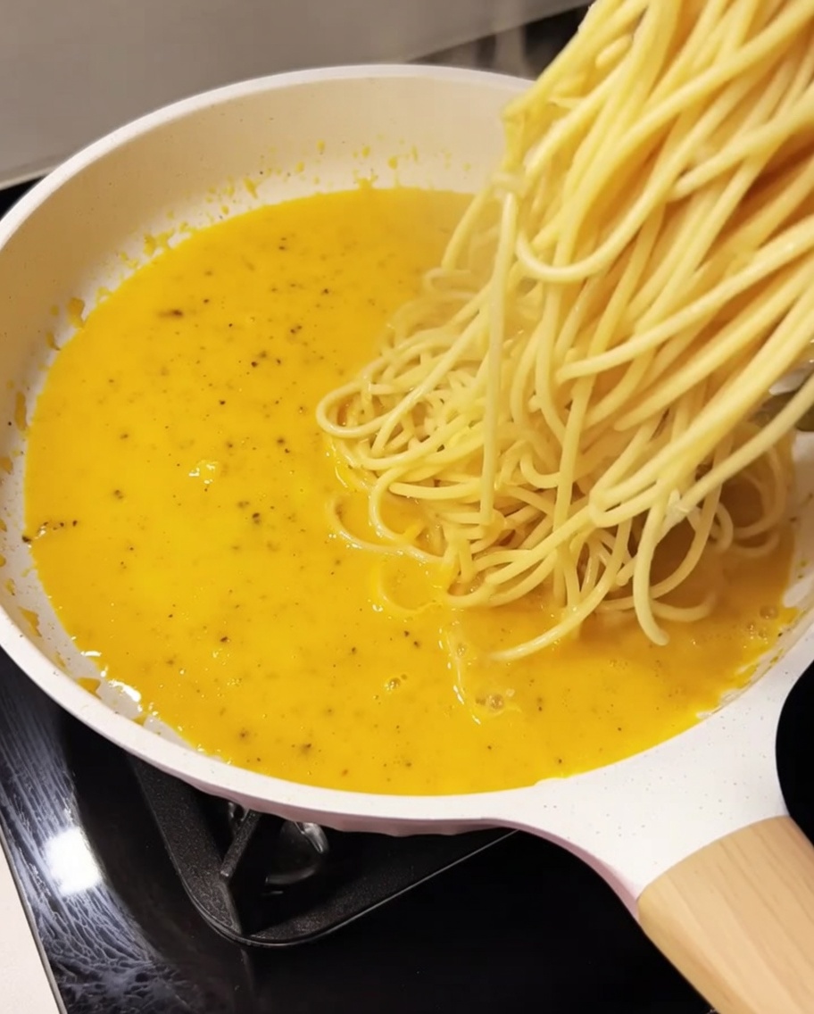 Cooked spaghetti being added to the creamy yellow sauce in a white pan.