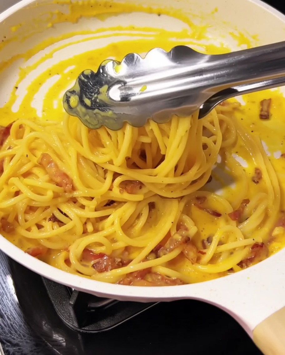 Tongs tossing spaghetti with sauce and guanciale in a pan.