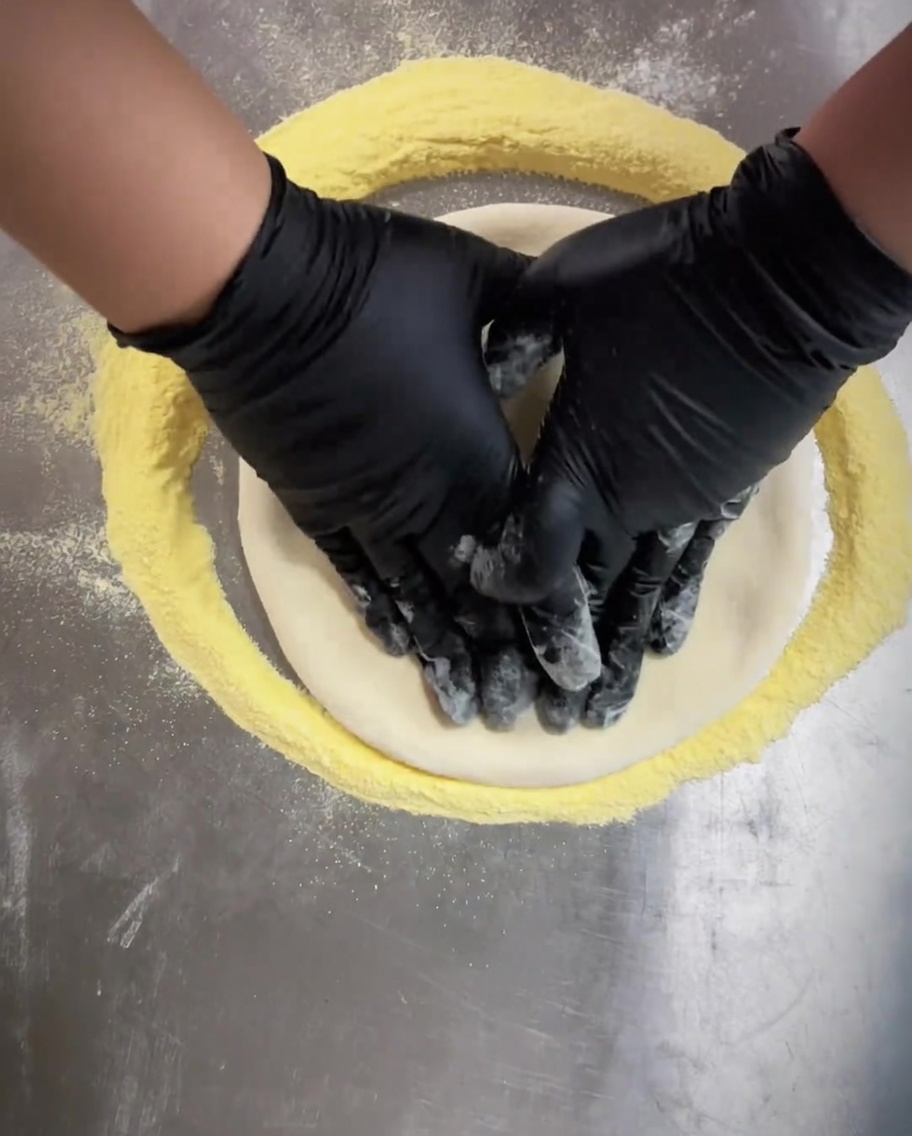 Hands wearing black gloves pressing into the center of the pizza dough to flatten it evenly.