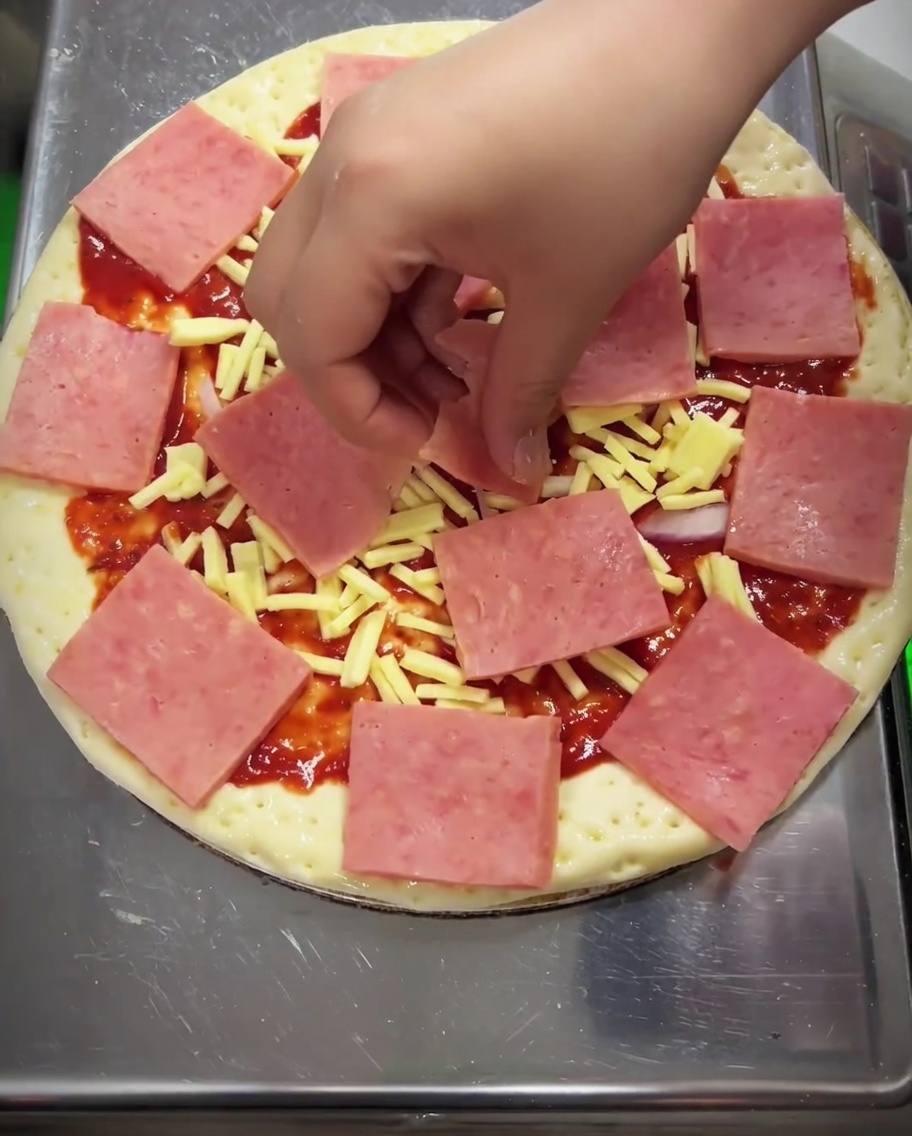 A close-up of a pizza being topped with yellow corn kernels, sliced green bell peppers, and pieces of mushrooms spread over the ham layer.
