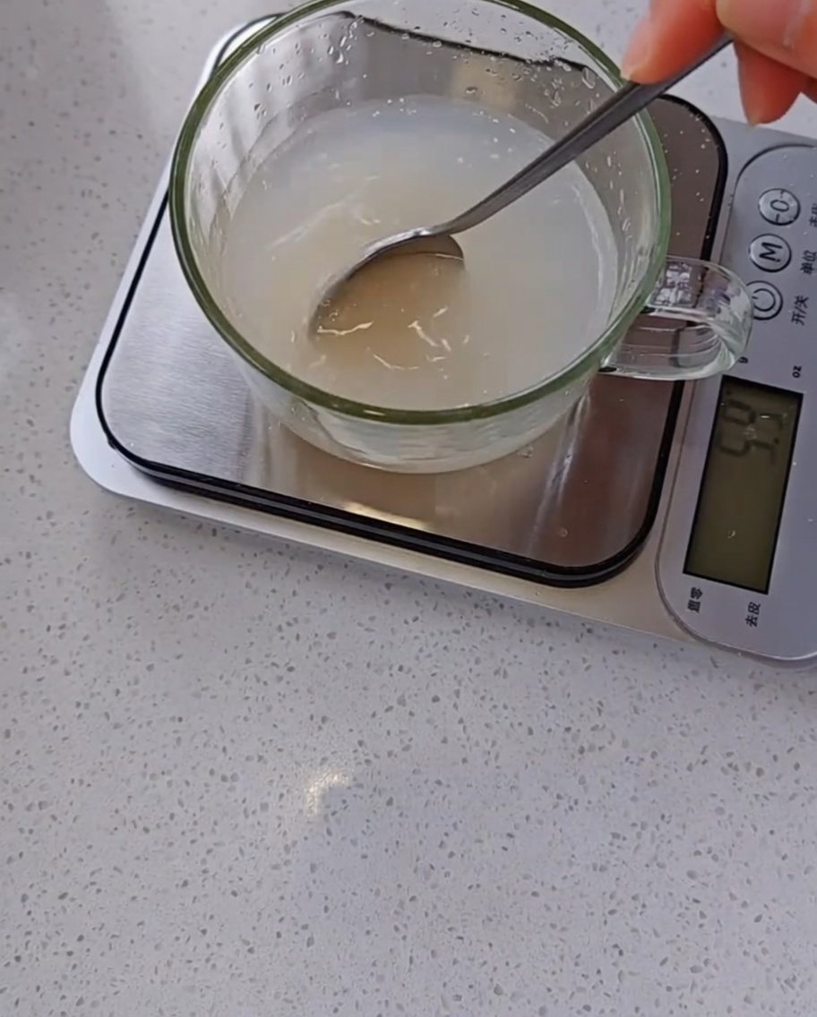A hand using a spoon to stir a cloudy mixture of yeast and water in a clear glass measuring cup sitting on a digital kitchen scale.
