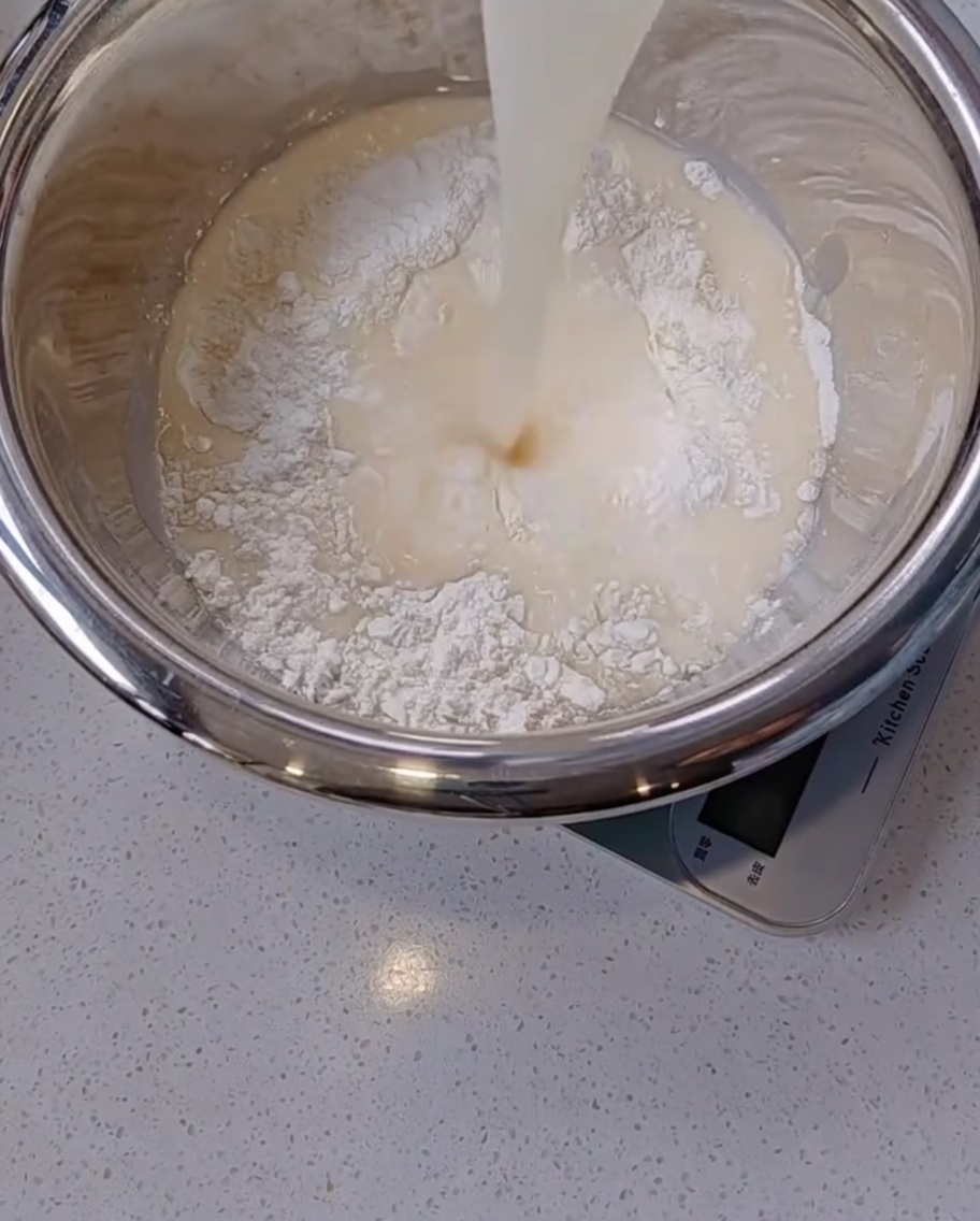 Pouring a milky yeast and water mixture from a glass cup into a stainless steel mixing bowl filled with dry white flour.