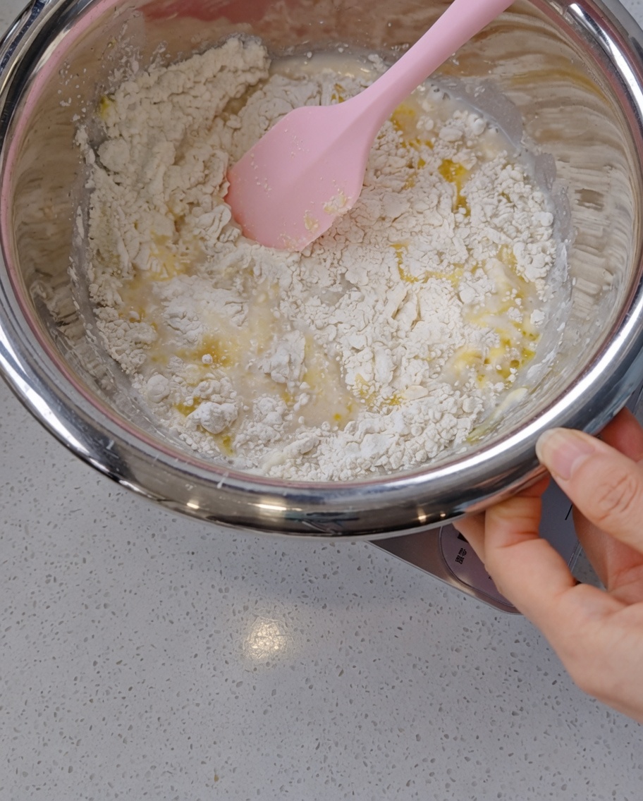A hand using a pink silicone spatula to mix a shaggy, wet dough with visible olive oil pools inside a stainless steel bowl.