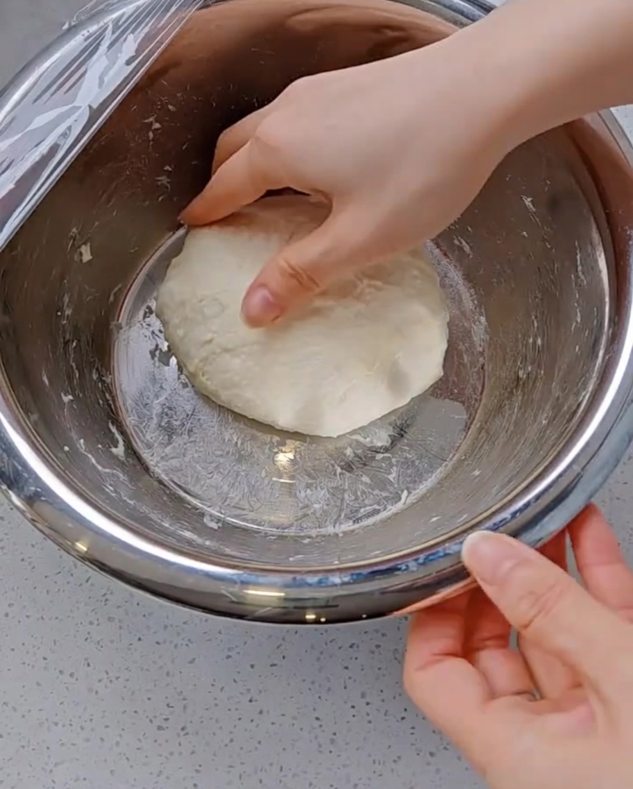 A hand stretching a piece of soft, elastic pizza dough upwards and folding it over towards the center of the smooth dough ball in a metal bowl.