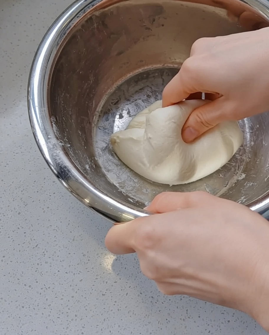 Hands gently tucking and rounding a smooth ball of pizza dough inside a stainless steel mixing bowl.