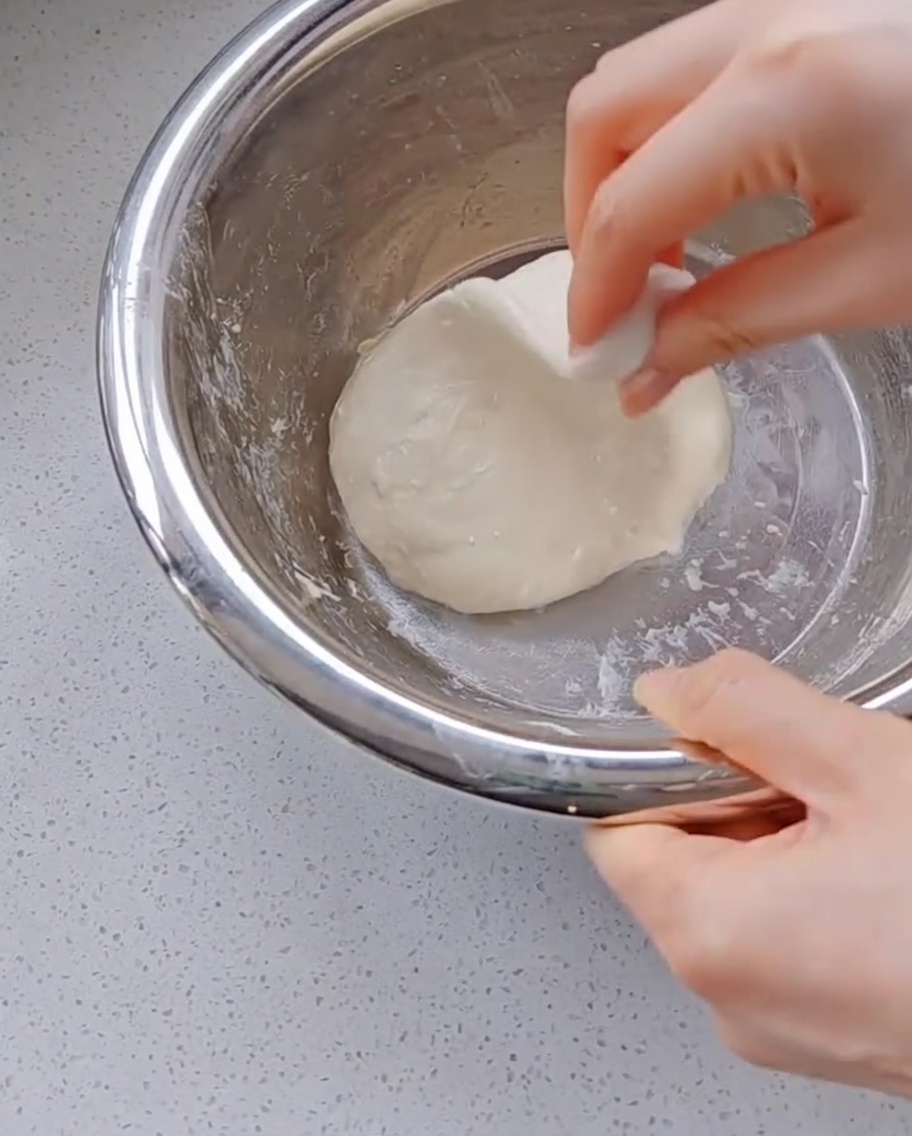 A hand pulling and stretching a piece of highly elastic pizza dough upwards inside a metal bowl.