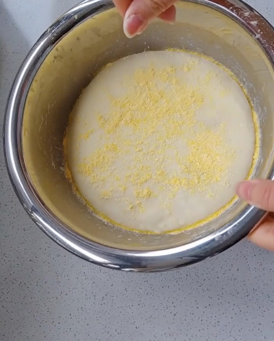 A hand sprinkling bright yellow cornmeal over a fully fermented, puffy ball of pizza dough inside a mixing bowl.