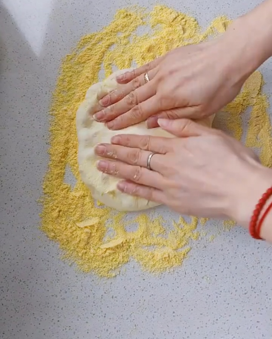 Hands using fingertips to carefully press and stretch pizza dough on a countertop generously dusted with yellow cornmeal.
