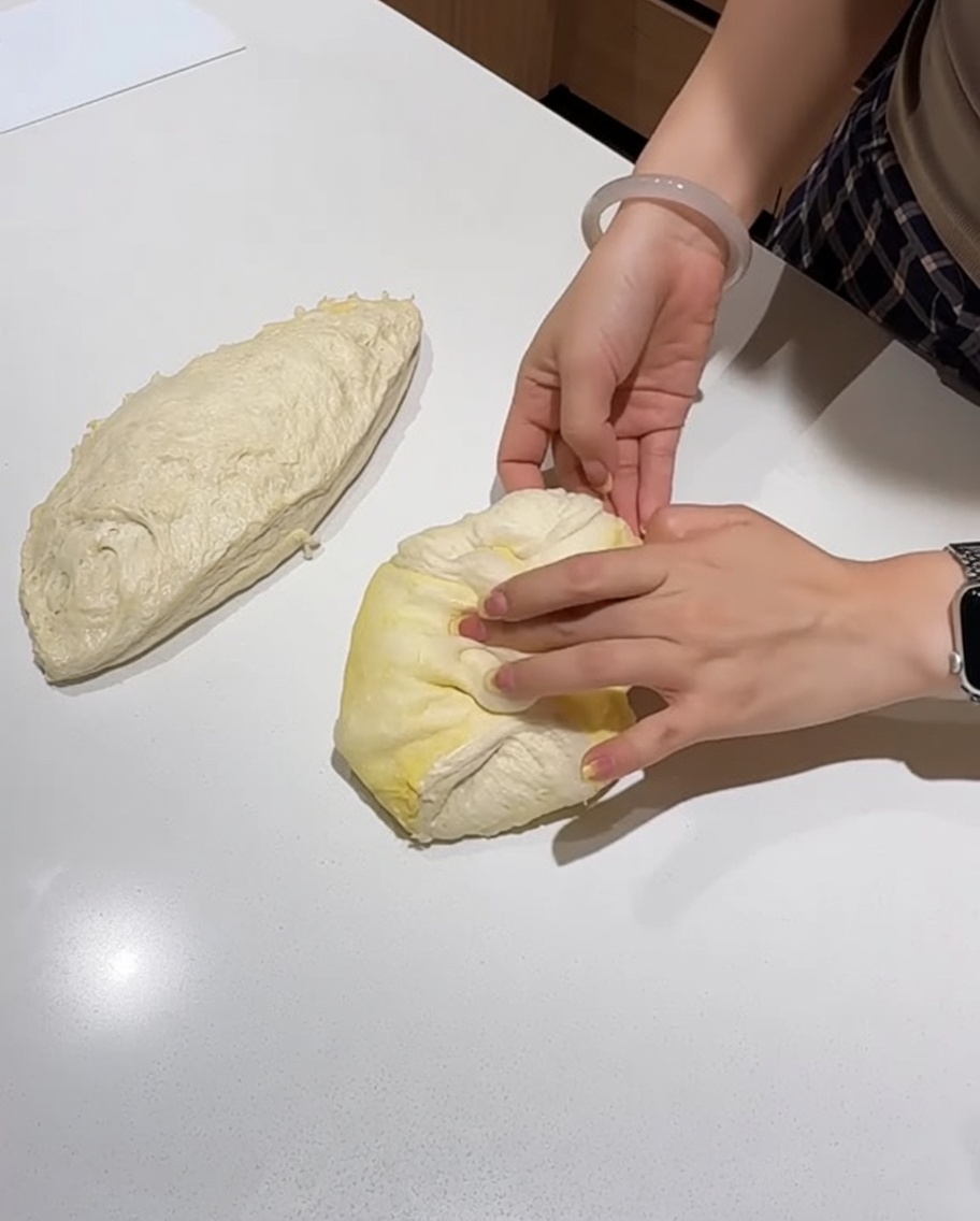 Hands folding and shaping a portion of pizza dough into a smooth, tight round on a white countertop.