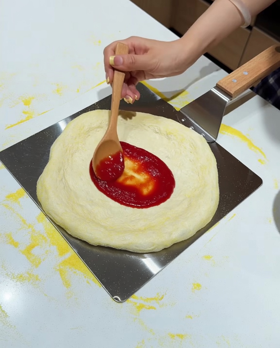A wooden spoon spreading red tomato sauce onto a stretched pizza dough resting on a metal peel.