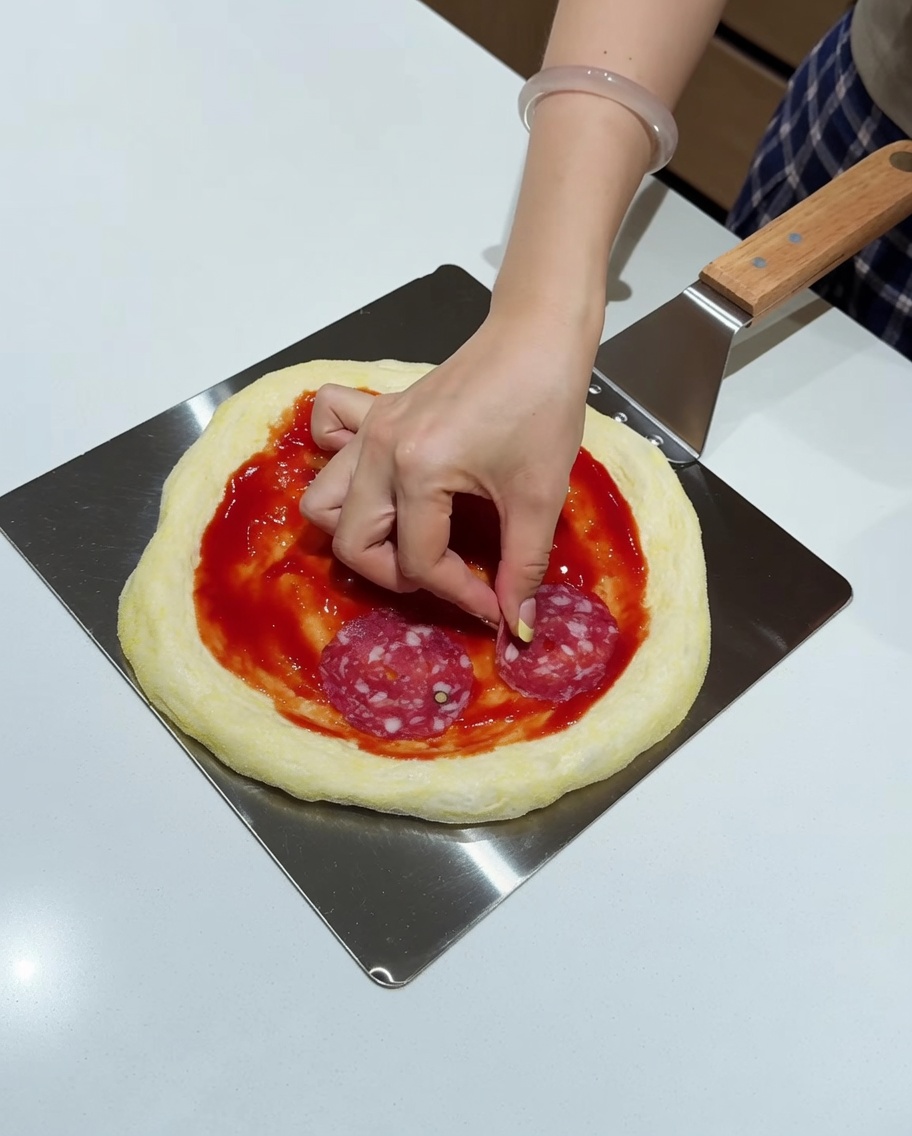 A hand placing round slices of salami onto a sauced pizza base on a metal peel.
