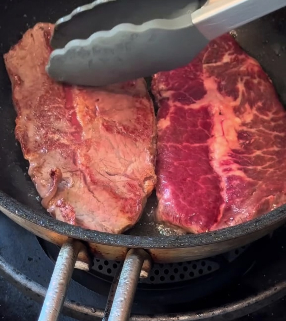 Two thin-cut steaks browning in a skillet with a fresh sprig of rosemary.